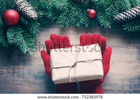 Female hands in red knitted gloves keep a Christmas gift against the background of a wooden old surface in an environment of fir-tree branches and Christmas tree decorations, top view, toned 