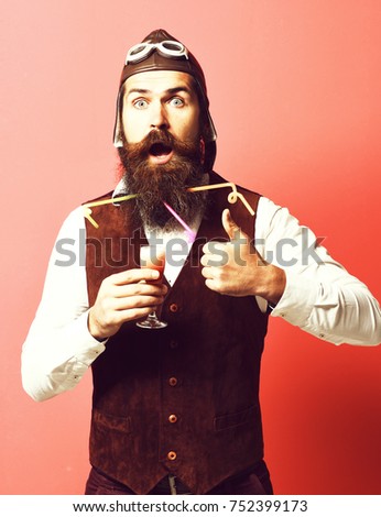 handsome bearded pilot or aviator man with long beard and mustache on surprised face holding glass of alcoholic shot in vintage suede leather waistcoat with hat and glasses on red studio background