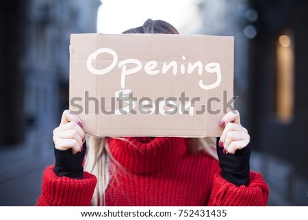 A cardboard tablet close-up with the inscription "Soon opening" in the hands of a young woman in a red sweater.