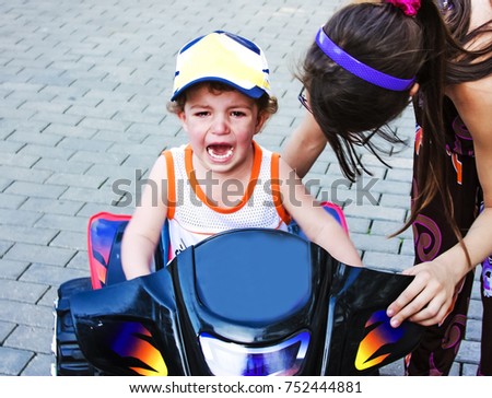 A little boy is capricious and crying, sitting on a children's ATV near the house. Children's games in the open air, walking and playing with a child on the street