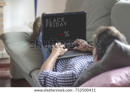 Young man lying on the sofa while using the laptop with Black Friday advertisement on screen. All screen graphics are made up.