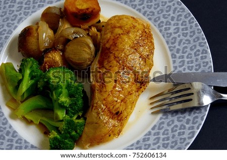 A piece of roast chicken breast is presented on a blue leopard patterned plate with roast vegetables and broccoli. A knife and fork complete the picture.