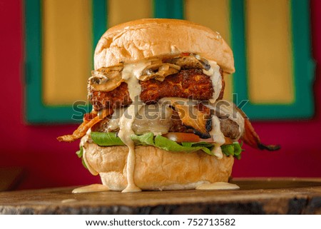 Delicious organic burger with mushrooms accompanied by fried cheese, lettuce, bacon, avocado and garlic sauce with spices, on a colorful background on a wooden table.
