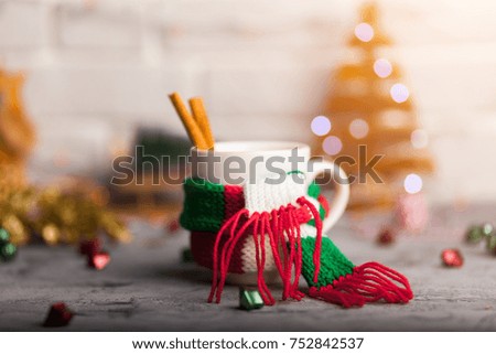 White cup of hot cocoa with mini marshmallows and cinnamon with green red white scarf against a rustic background with beautiful Christmas lights of bokeh with toys