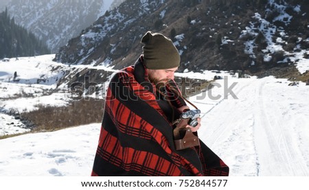 Hipster bearded man in red blanket looking in retro film camera. Photographer in mountains in cold sunny winter day