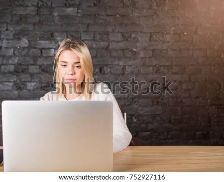 Confident woman developer sites working on laptop computer, sitting in enterprise office against brick wall with copy space. Blonde female reading financial news in internet via portable net-book 