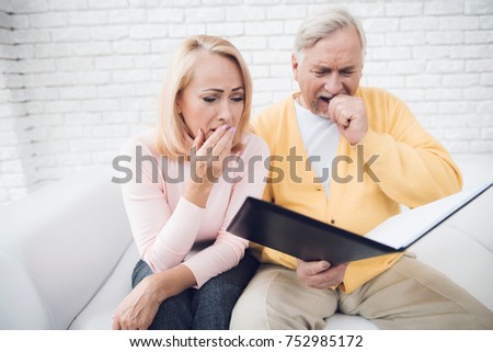 A man in a yellow cardigan and a woman in a pink sweater are sitting and reading documents in a black folder. They have a shock on their face, they are very upset.