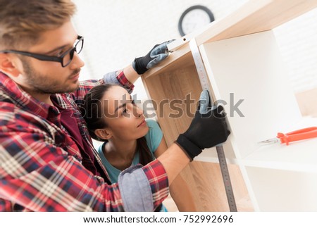 A man and a girl add up a bookshelf for their apartment. The bookcase is made of wood. It is added by bolts. They measure the boards with a ruler.