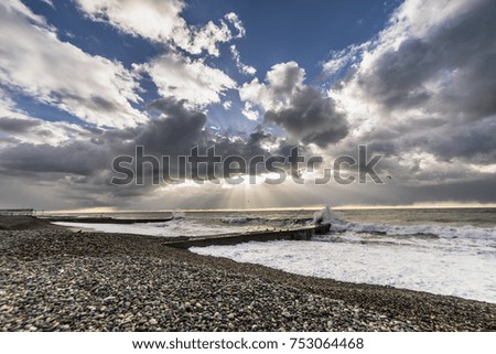 the beach from the pebbles rages the sea, white foam from the waves, the sun is covered by gray clouds