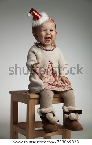 excited little girl wearing santa hat is screaming while sitting on a chair on grey background