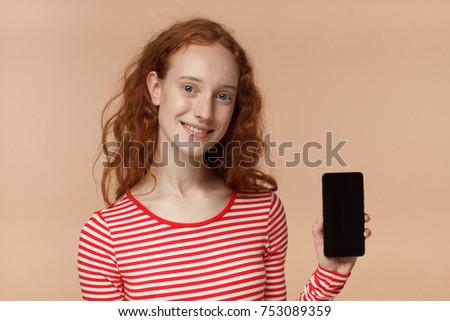 Indoor closeup of beautiful redhead teen girl isolated on peach background showing blank phone screen with copyspace for advertisement of your services or goods, concept of advice and recommendation