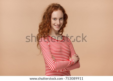 Horizontal indoor photo of good-looking redhead teen girl isolated on peach background dressed in casual clothes, standing with crossed arms and cheerful face expression, being joyful and calm