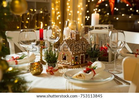 Beautiful served table with decorations, candles and lanterns. Little gingerbread house with glaze on white tablecloth. Living room decorated with lights and Christmas tree. Holiday setting Royalty-Free Stock Photo #753113845