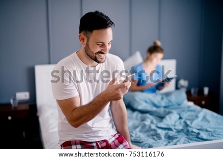 Smiling satisfied handsome husband reading a message in front of a girl with a tablet on the bed.
