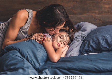 Pretty little girl is sleeping in bed at home, mom is kissing her in cheek