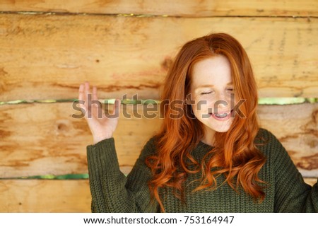 Young cheerful red-haired woman with eyes closed standing against wooden fence
