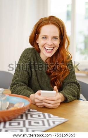 Portrait of young cheerful red-haired woman sitting at table with smartphone