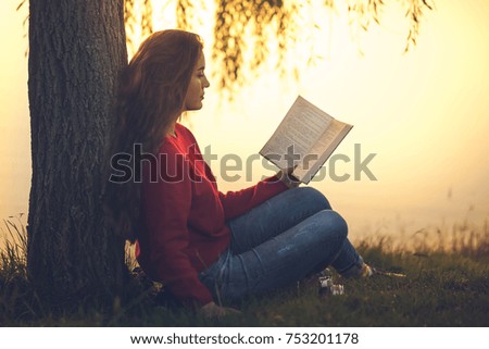 Beautiful young woman sitting and reading book in a park near the lake.