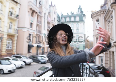 A stylish and positive girl smiles and takes a sephi on the background of a beautiful old town. A tourist woman is photographed on her phone while walking