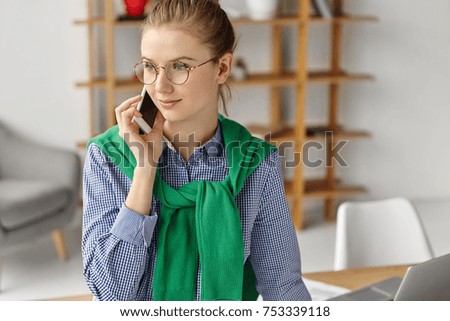 Creative female designer sits at her cabinet, looks pensively aside, calls family to say that she is late from work, has much blueprints to creat. Woman in eyewear communicate on smart phone in office