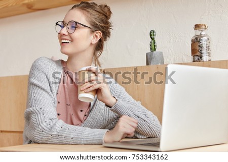 Carefree female writer works at new novel or detective story, keyboards on laptop, looks with smile aside, glat to notice best friend, drinks hot coffee from takeaway paper cup, enjoys free wifi