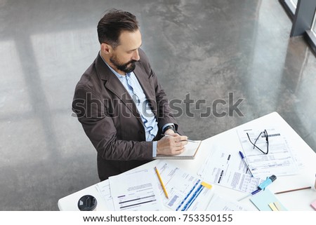 Top view of handsome skilled professional employer dressed formally, sits at work place, surrounded with many documents, writes with pencil in papers. Male marketing specialist works indoors