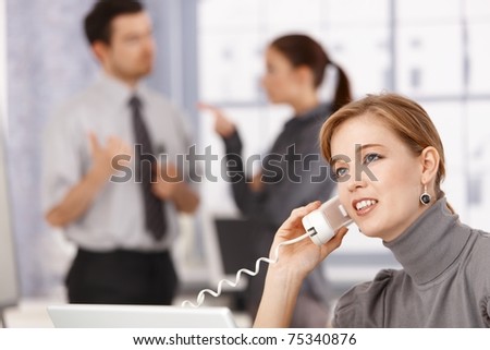Young businesswoman talking on phone in office, colleagues chatting in background.?