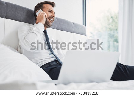 Laughing businessman talking on phone while sitting on bed with laptop