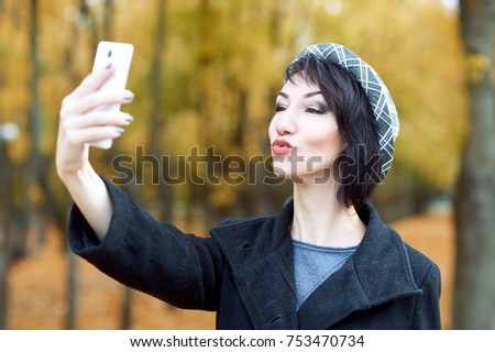 girl photographs herself on the phone and kisses in autumn city park, yellow leaves and trees, fall season