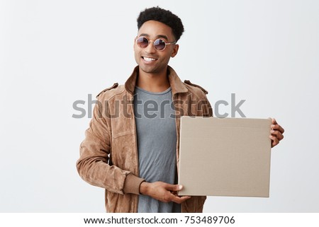 Present for you. Isolated on white portrait of young fashionable dark-skinned man with afro hairstyle in grey t-shirt, brown jacket and sun-glasses holding box in hand, smiling in camera