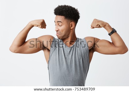 Close up of young athletic handsome dark-skinned man with afro hairstyle in sporty grey shirt looking at his muscles with concentrated and confident face expression. Healthy lifestyle.