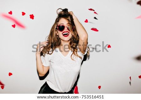 Indoor portrait of amazing caucasian female model in trendy t-shirt touching her long shiny hair. Laughing refined woman in sunglasses having fun with red confetti. Royalty-Free Stock Photo #753516451
