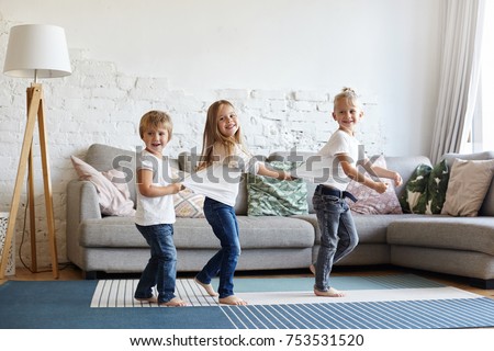 Picture of three excited children siblings in white t-shirts and jeansdancing barefooted on wooden floor at home. Two little girls standing in sequence with their blonde elder brother in the lead
