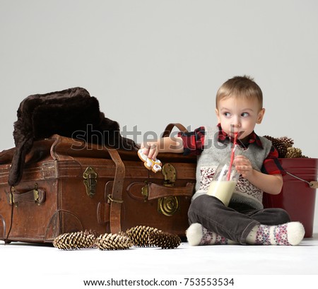 Small child baby boy in winter hat sitting near retro leather travel bag in winter snow hat  drinking bottle of milk. Christmas concept on grey background