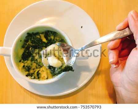 Spinach with cheese in white cup and plate on wood table with woman’s hand holding spoon is scooping it. It is Italian food. Concept for restaurant business with space for text design. Blurry picture.