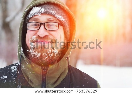 Closeup portrait of happy young bearded man in cold weather in the winter forest at sunset. Fun guy smiles. Face, beard , mustache, and glasses misted, covered with frost.
