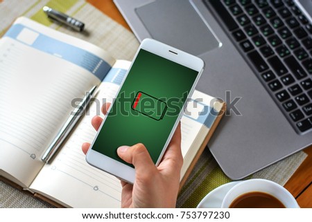 A woman's hand holds a smartphone with low battery sign over a notebook, a cup of coffee and a laptop