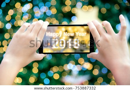 Girl's hand holding mobile phone take a photo of christmas tree with wording Happy New Year 2018. Young woman using smartphone outdoor capture picture of bokeh blur Christmas lighting lamp decoration.