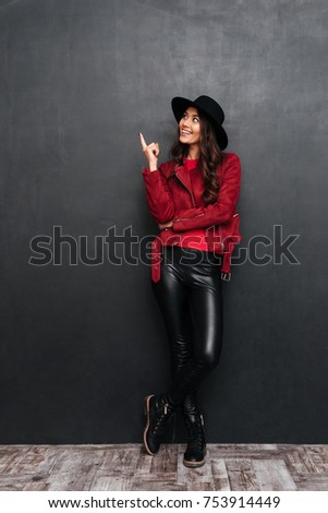 Picture of happy young beautiful woman wearing hat standing over dark grey wall chalkboard pointing. Looking aside.