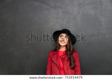 Picture of happy young woman wearing hat standing over dark grey wall chalkboard. Looking aside.