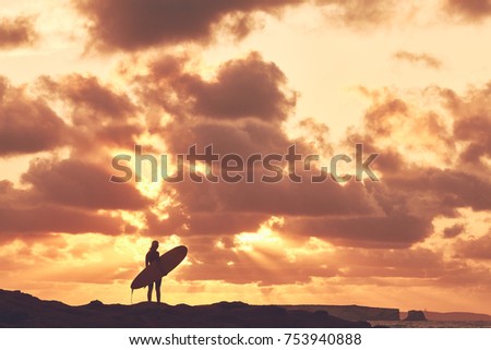 Young woman wearing shorty wetsuit, standing on beach with surfboard in her hands and enjoying stunning sunset after evening surfing session - water sports concept. Baleal, Peniche, Portugal