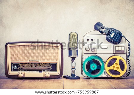 Retro broadcast radio receiver, reel to reel tape recorder circa 60s, microphone and headphones on wooden table front concrete wall background. Vintage style filtered photo
