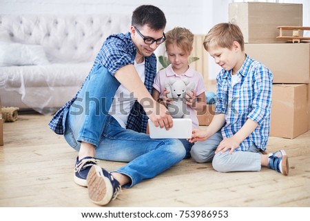 Lovely moments of fatherhood: handsome young dad sitting on floor of living room with his cute little children and watching cartoon on digital tablet, pile of moving boxes behind them