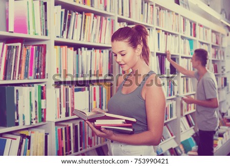 portrait of  happy european teenage girl customer looking at open book standing among bookshelves 