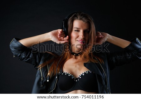 Studio photo of beautiful model in headphones with hands at head, underwear and leather jacket listening to music on empty black background