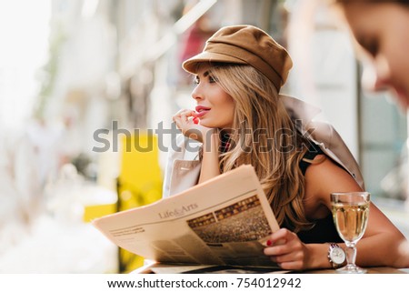 Dreamy woman in brown cap thinking about something, propping face with hand and holding newspaper. Outdoor portrait of amazing blonde girl with glass of wine posing on blur city background.