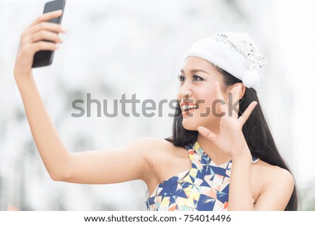 Beautiful toothy smiling young woman in white santa's hat  using smartphone doing facetime with family against blur background.Christmas concept.