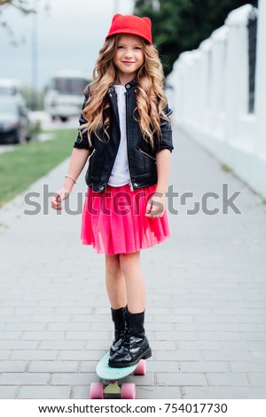 Stylish fashion little girl child riding skateboard on city street. Red hat, black biker jacket, red skirt, white T-shirt and boots. Happy child of a teenager. Beautiful long hair.