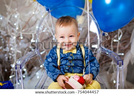 A handsome boy sits on a transparent chair. In a blue shirt and yellow bars with a yellow butterfly. In the background are balloons.