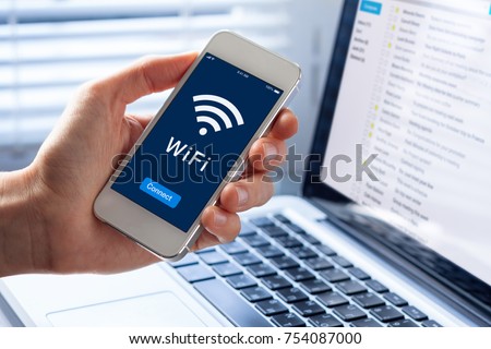WiFi symbol on smartphone screen with button to connect to wireless internet, close-up of hand holding mobile phone, computer in background Royalty-Free Stock Photo #754087000